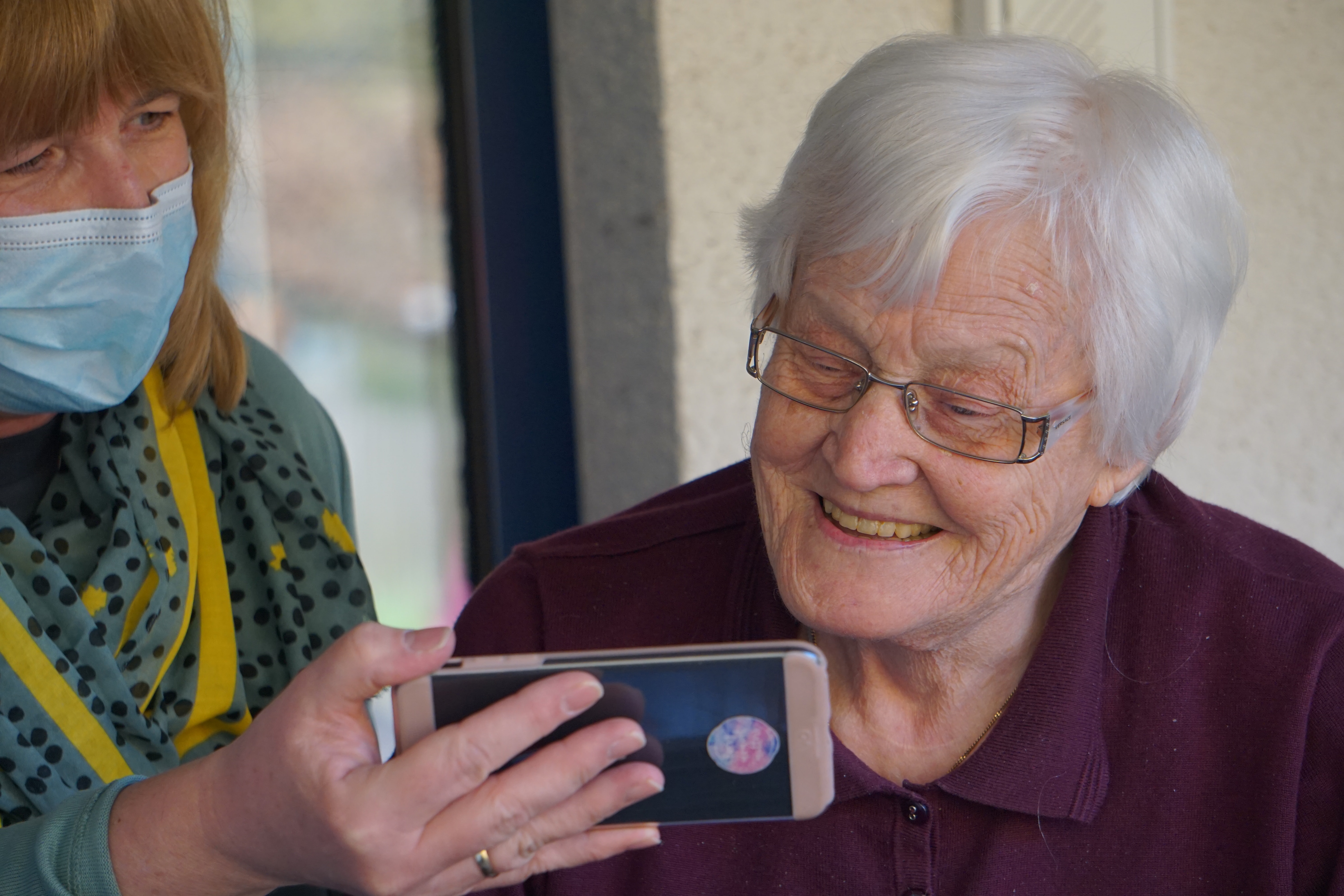 Elderly woman and carer wearing a face mask looking at a smart phone. 