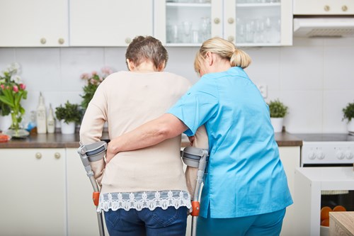 A health care worker supporting a woman using crutches to walk in her kitchen.
