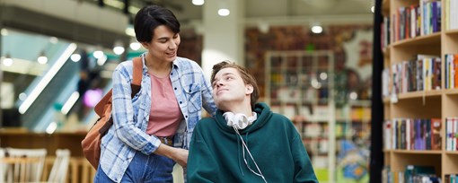 Two young adults in a library. One is supporting the other who is in a wheelchair. 